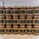 Row of glass jars with gold lids labeled 'Moutarde en grains' on a shelf, containing whole-grain mustard.