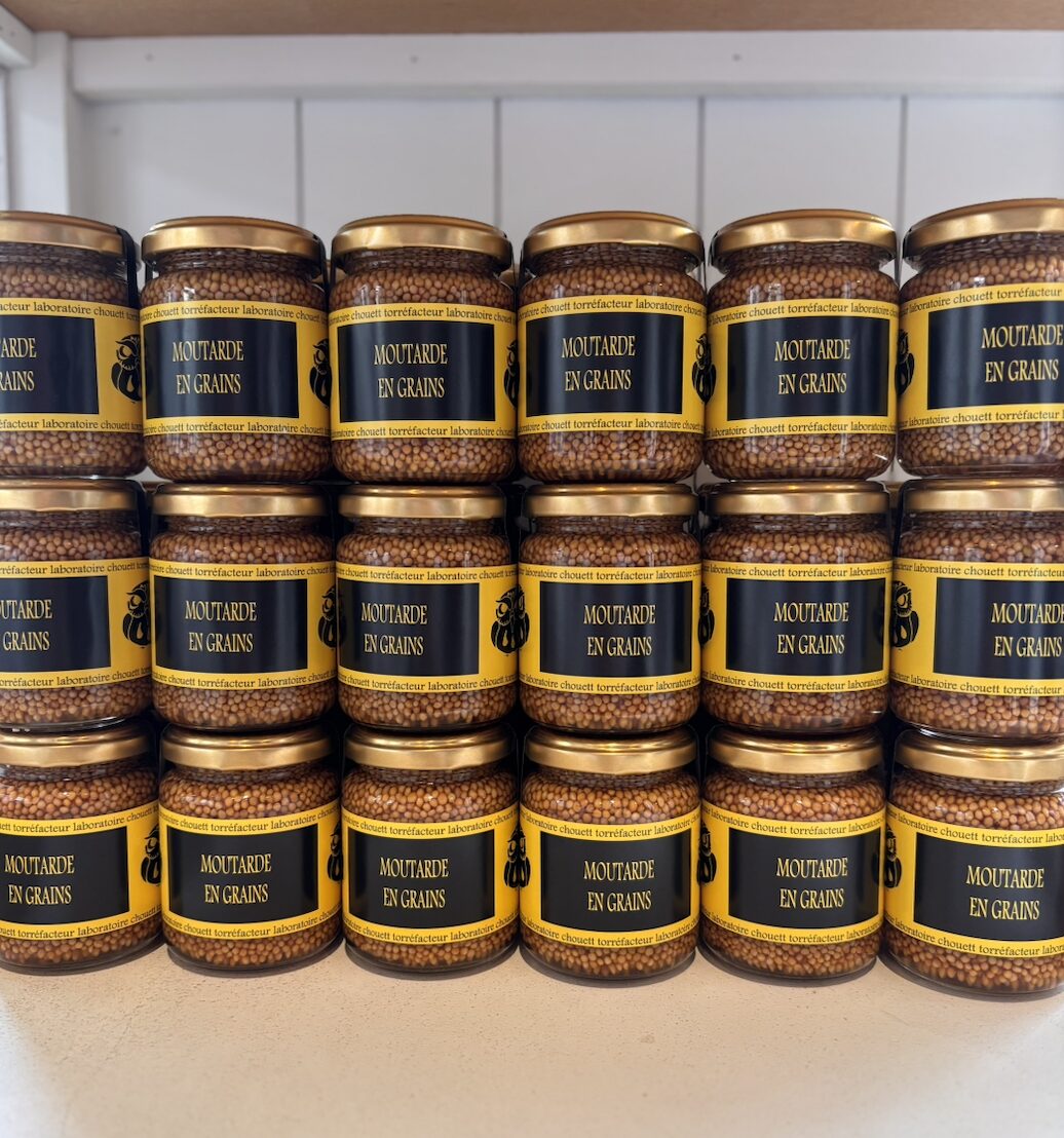 Row of glass jars with gold lids labeled 'Moutarde en grains' on a shelf, containing whole-grain mustard.