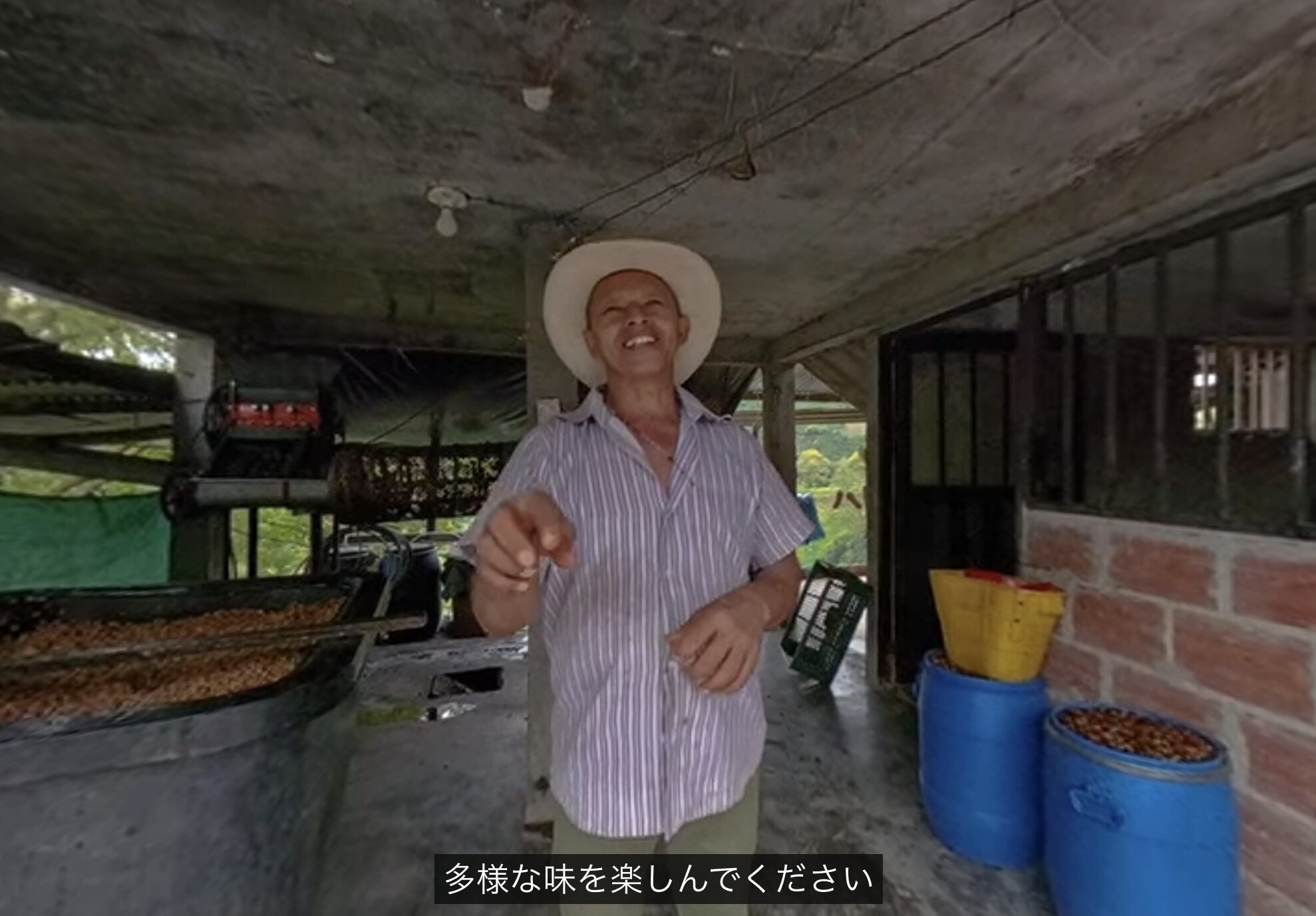 スクリーンショット 2026-07-06 112334 | chouette torréfacteur laboratoire Smiling man in a striped shirt and white cowboy hat points toward the camera inside a covered market stall and crates behind him.
